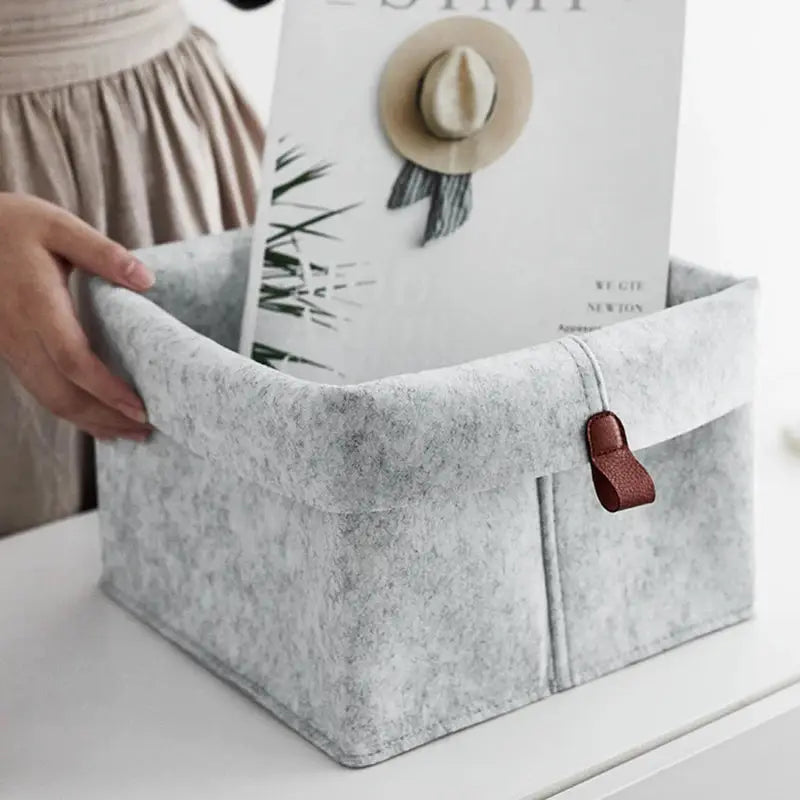 Grey felt storage basket for clothes with brown leather strap, a compact home organizer.