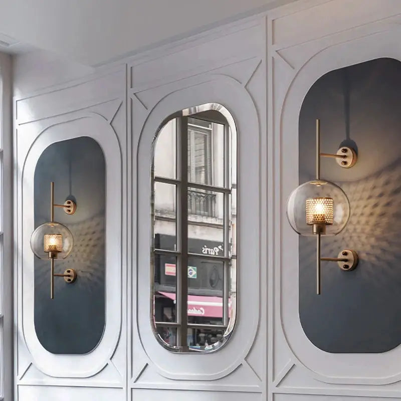 Pair of modern glass wall lamp sconces with gold metal arms and spherical glass shades on a decorative panel wall.