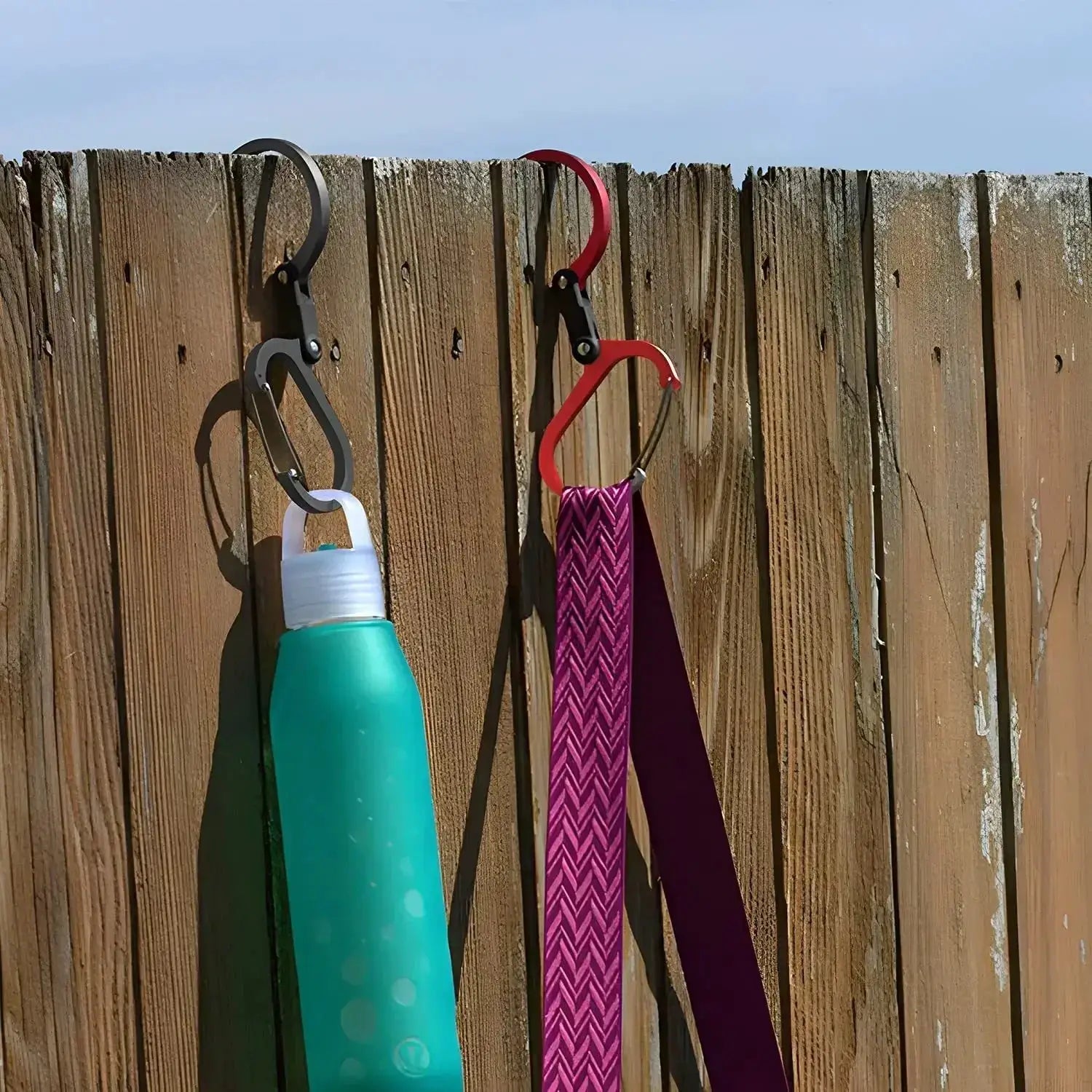 Two rotating carabiners (black and red) attached to a wooden fence, with a water bottle and strap.