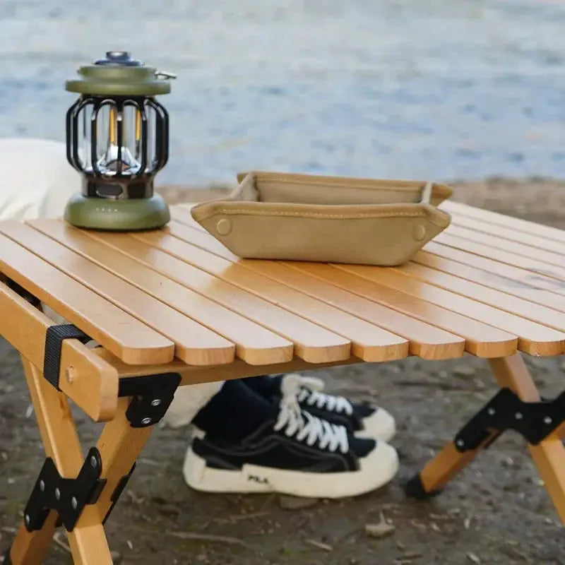 Portable camping organizer tray in tan fabric on a wooden foldable table beside a green lantern.