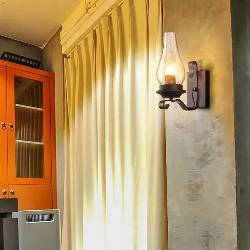 Wrought iron wall lamp with glass hurricane shade in black finish, mounted beside beige curtains.