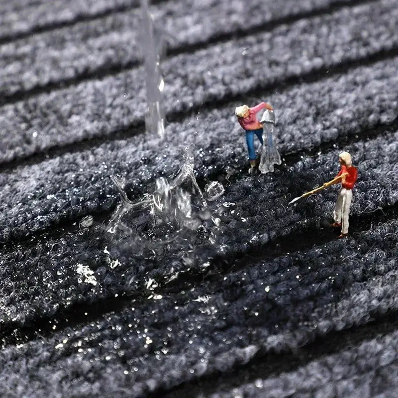 Door mat in dark black fibers; water-absorbent and anti-slip, suitable for indoor or outdoor use with two tiny figurines for scale