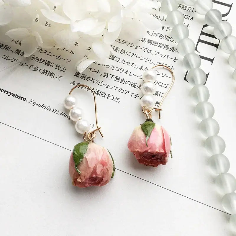 Pair of rose drop earrings with pink dried-rose pendants encased in epoxy resin, gold-tone hooks, and pearl beads.