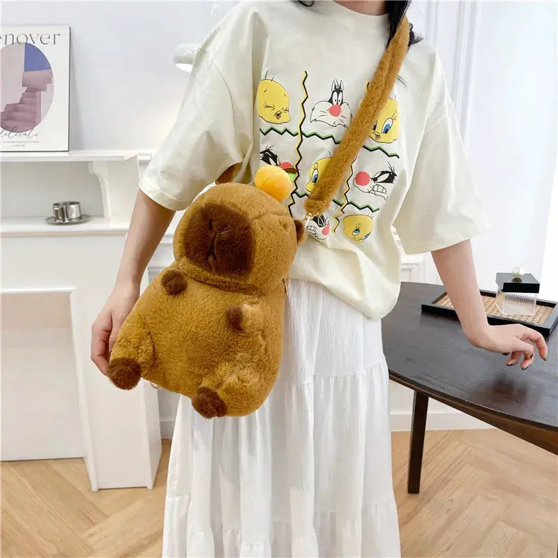 Capybara plush backpack in brown fuzzy fabric with tiny ears, worn as a crossbody bag by a girl in a beige tee.