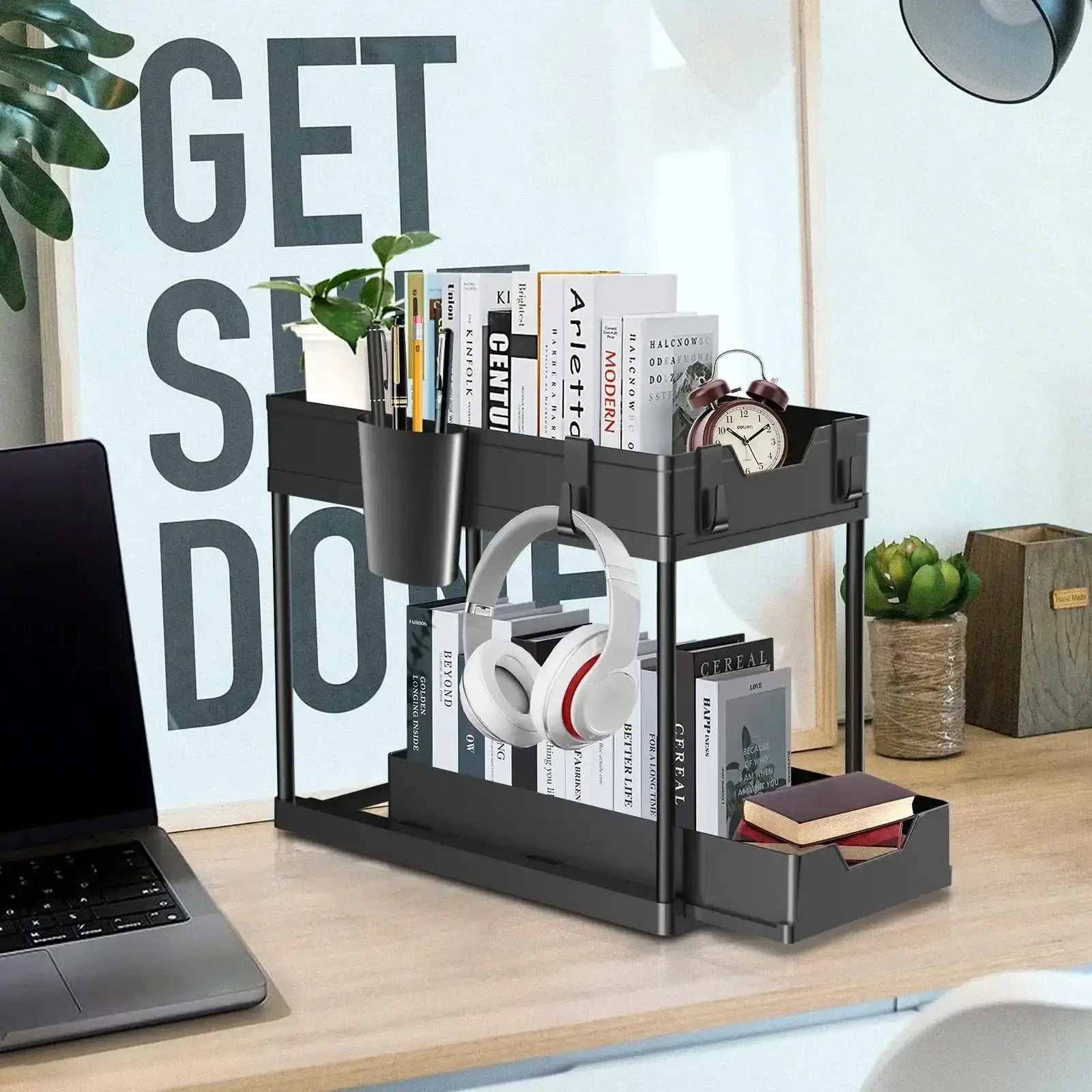 Two-tier black under-sink sliding storage rack with hooks; holds books, headphones, mug and desk items.