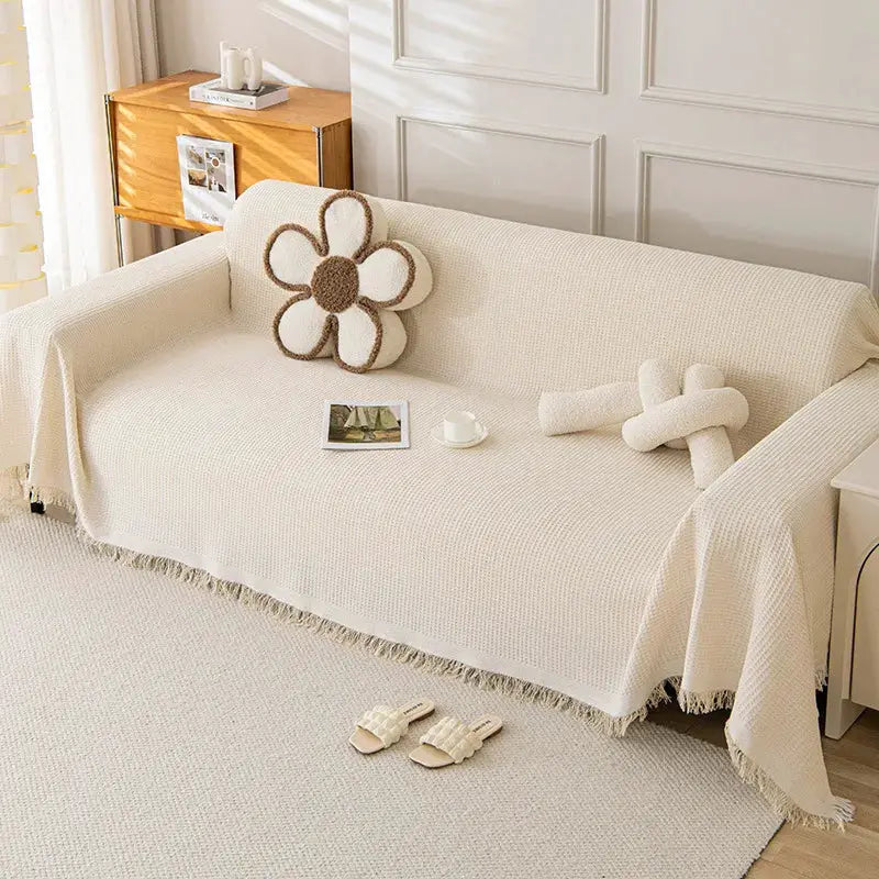 Sofa towel cover by Simples Market in cream knit with fringe; shown with one flower cushion and two rolled towels.