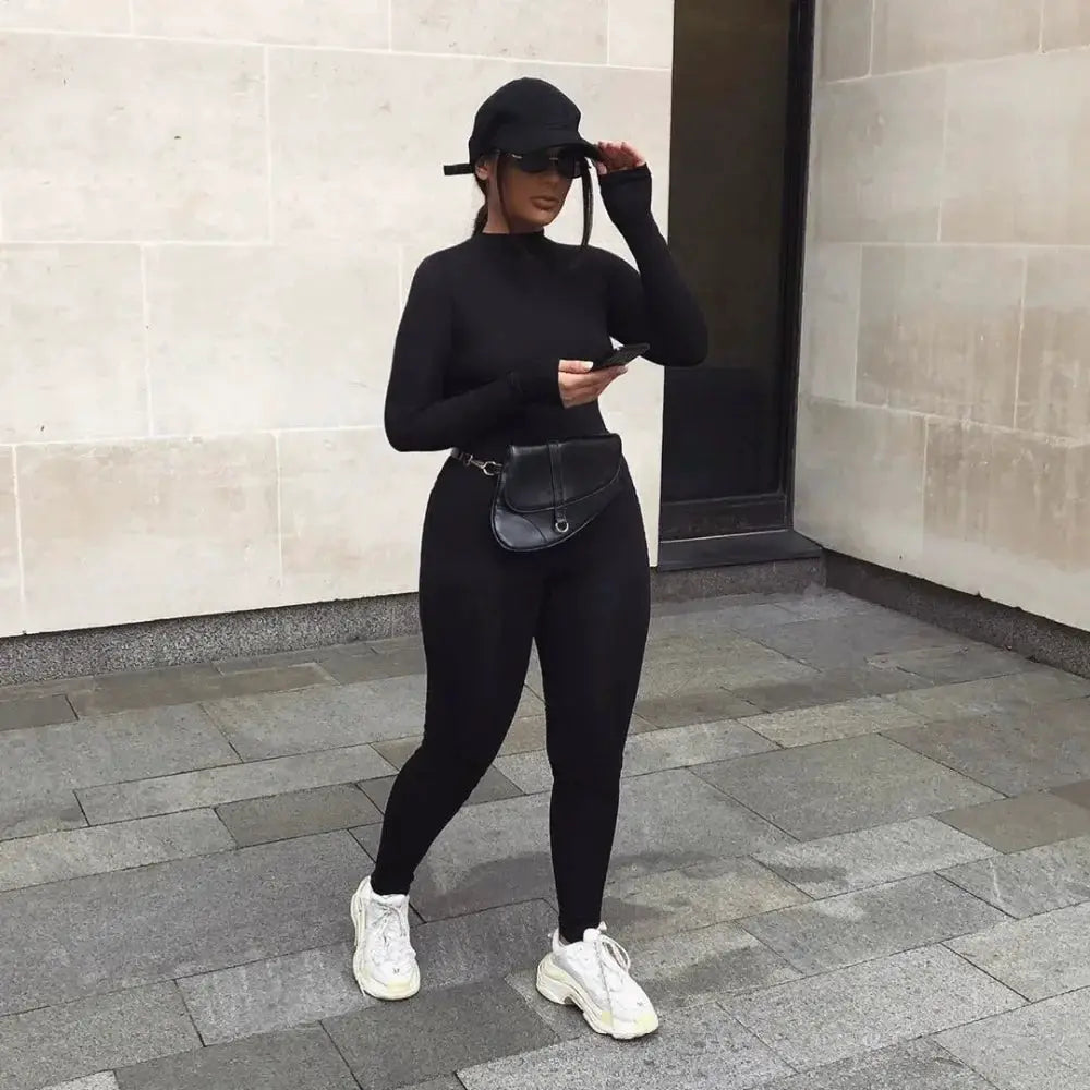 Black long-sleeve jumpsuit for women, stretchy and form-fitting, with a black waist bag and white sneakers.