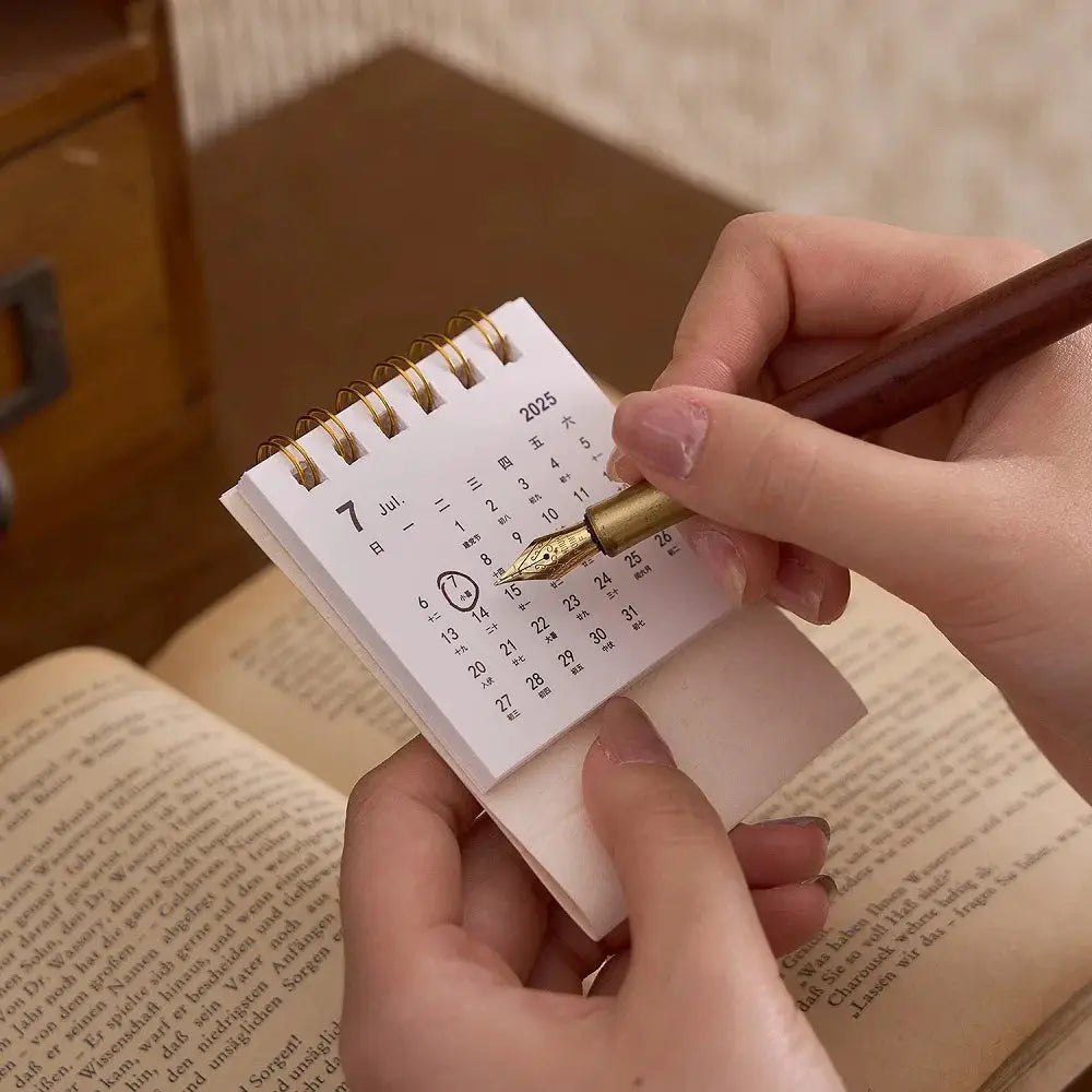 Mini desk calendar with brass coil binding and white pages, being written with a fountain pen.