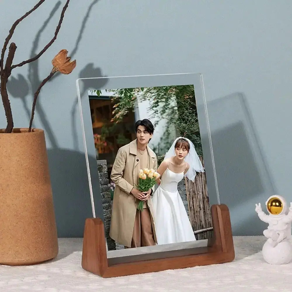Acrylic U-shaped photo frame with walnut/beech wooden base on desktop, displaying a wedding photo