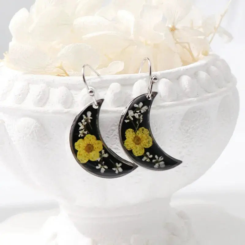Pair of crescent moon pressed-flower epoxy resin earrings by Simples Market with yellow blooms on black