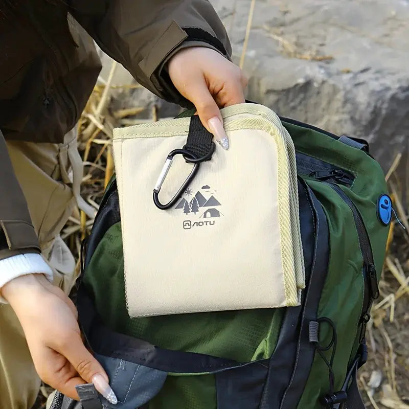 Outdoor camping foldable seat cushion in cream fabric, moisture-proof mat, attached to green hiking backpack.