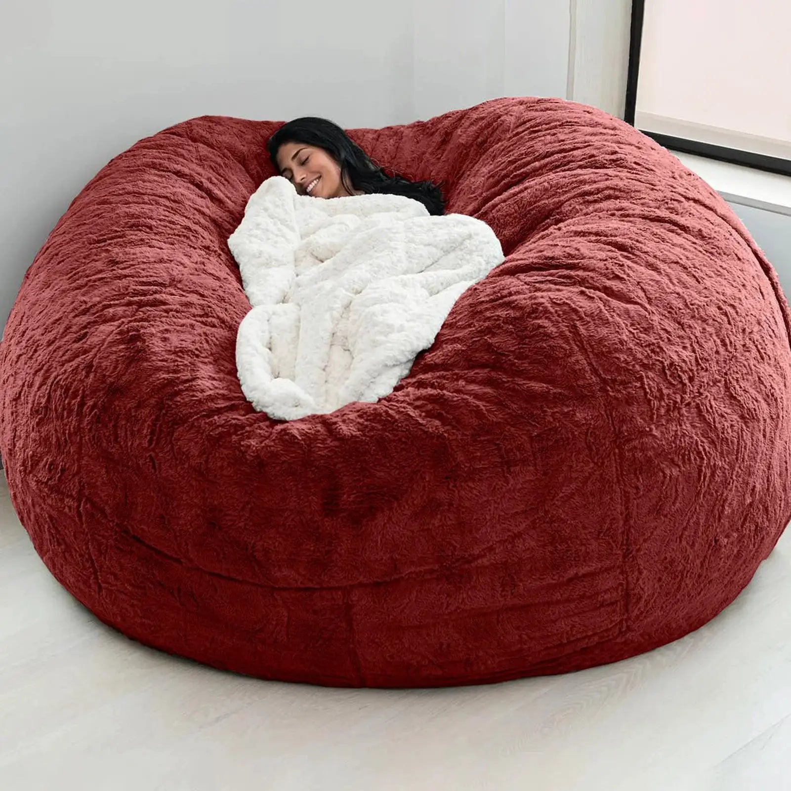 Bean bag sofa bed in red plush fabric with a white fluffy blanket, person relaxing.