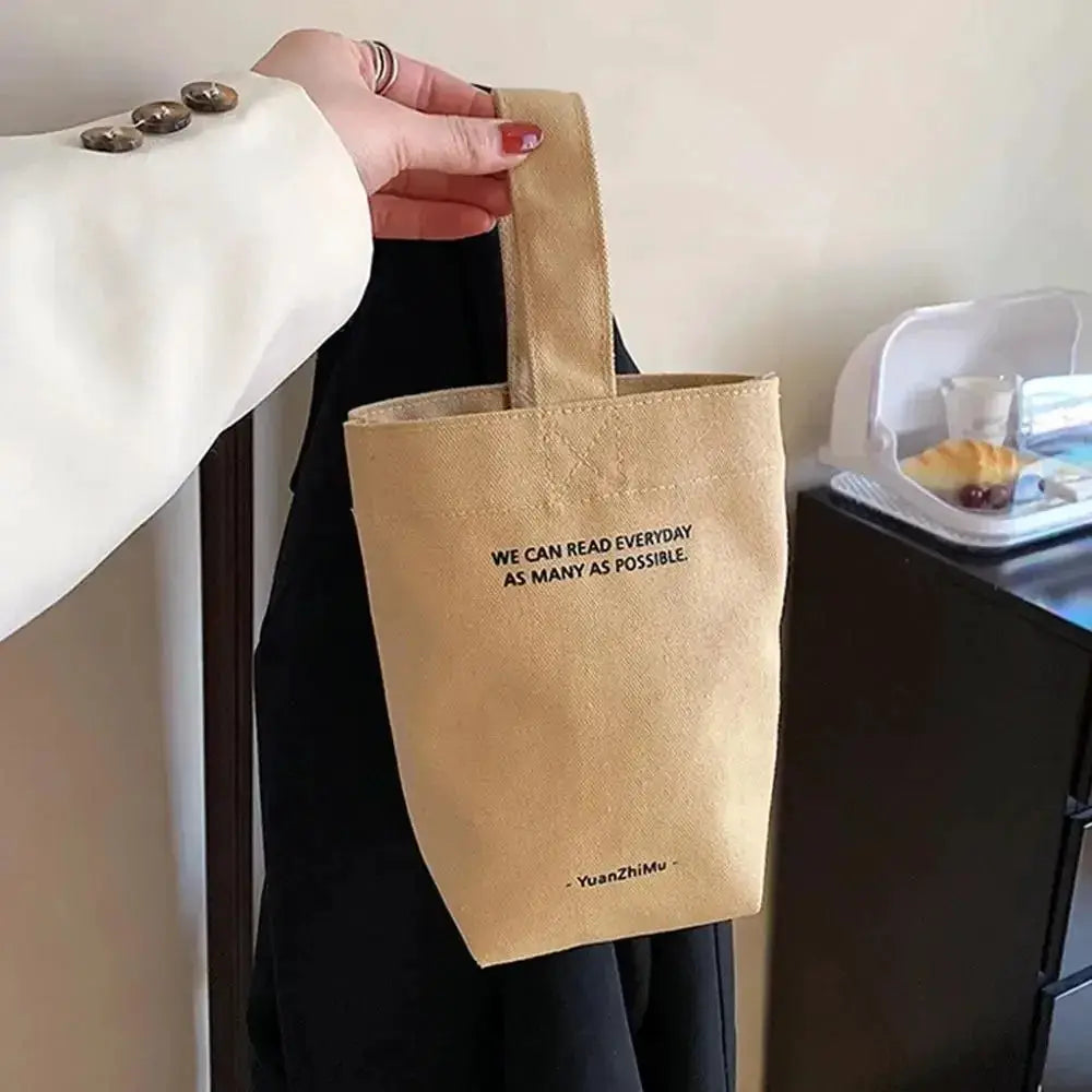 Beige canvas shoulder bag with short handle; large-capacity tote featuring black slogan text.