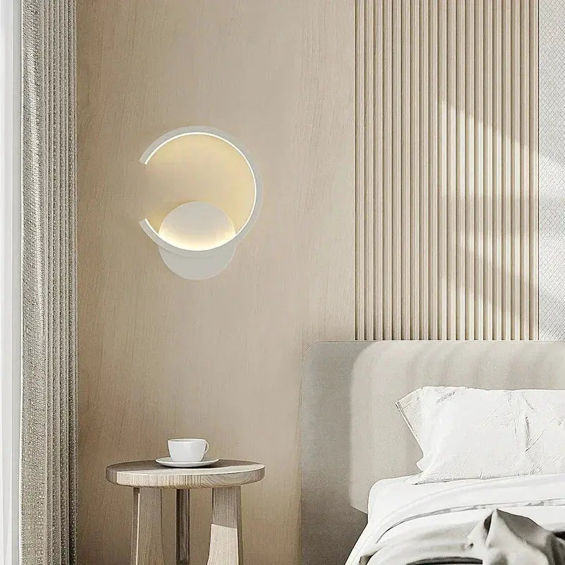Modern LED wall lamp, white circular ring with warm gold glow, mounted on light wood wall beside bed