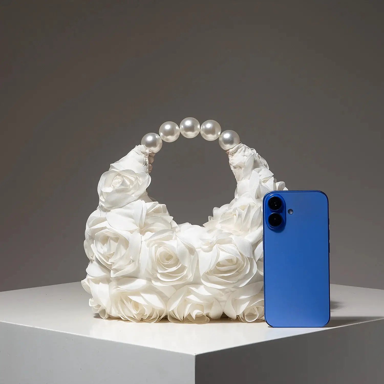 Cartera para mujer elegante con asa de perlas y flores blancas, junto a un teléfono azul
