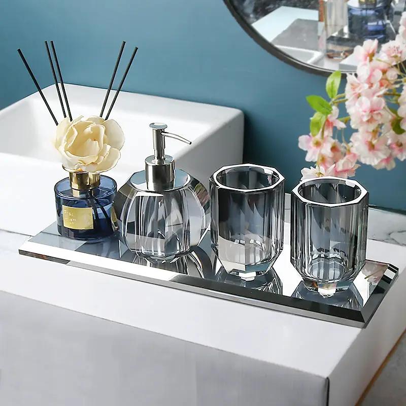 Four-piece glass bathroom set on a mirrored tray: diffuser bottle with reeds, pump dispenser, and two smoky glass cups