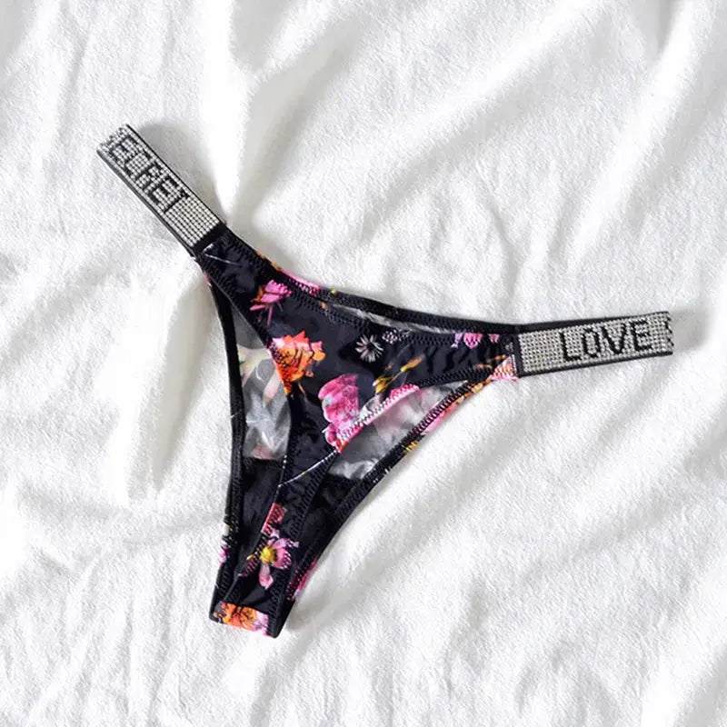Thong underwear, low-waist floral print with rhinestone LOVE waistband on black fabric.