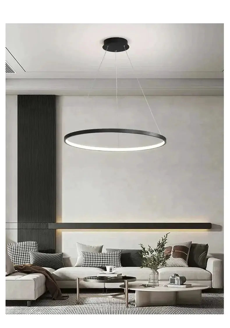 Modern round LED pendant light with black metal ring and white glow, suspended by cables over a living room.