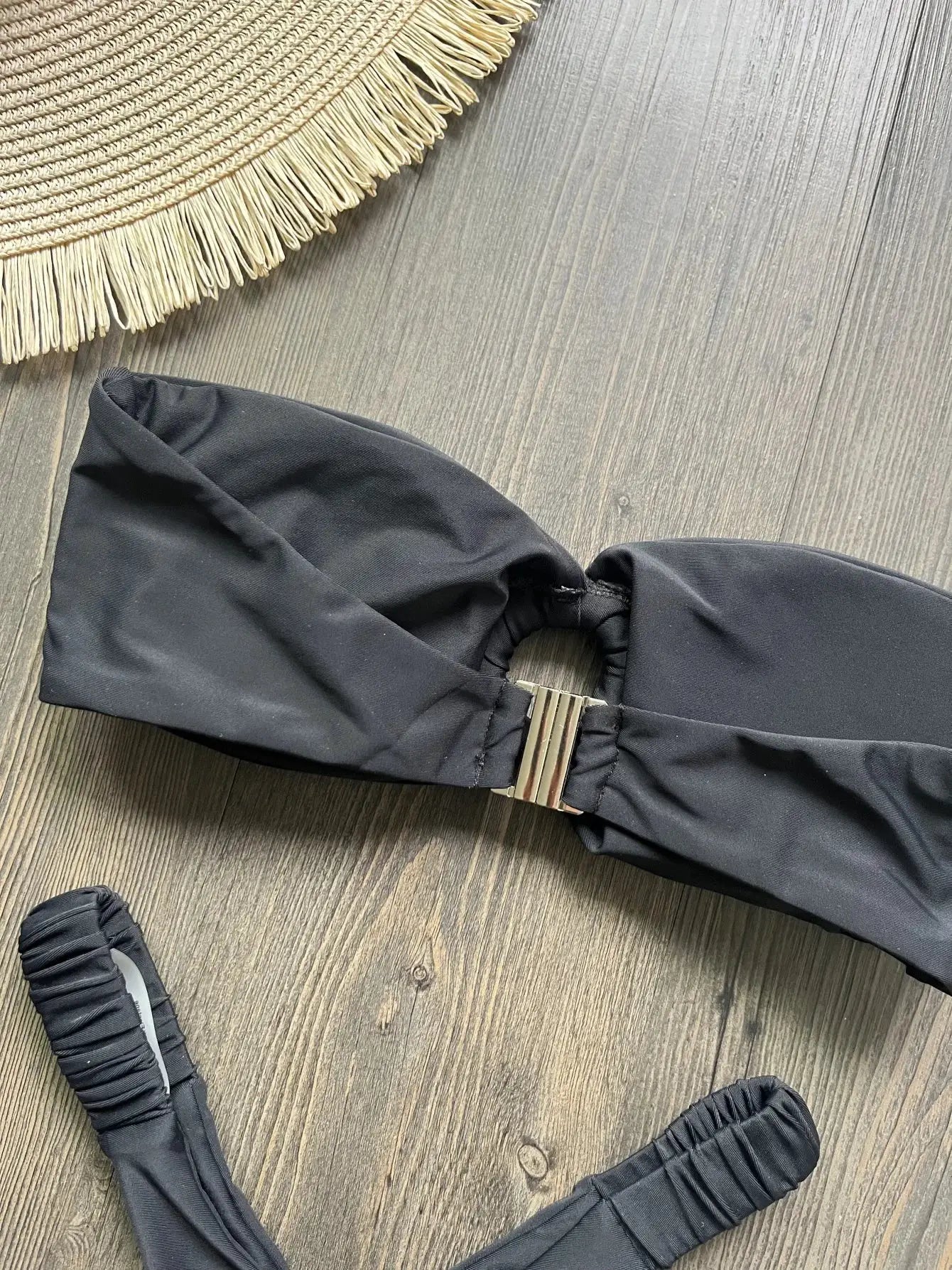 Black micro bikini top with gold clasp, push-up styling, on wooden surface beside a straw fringe hat