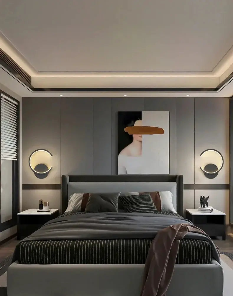 Modern LED wall lamp pair with circular halo design; black frames and warm lighting beside a gray upholstered bed.