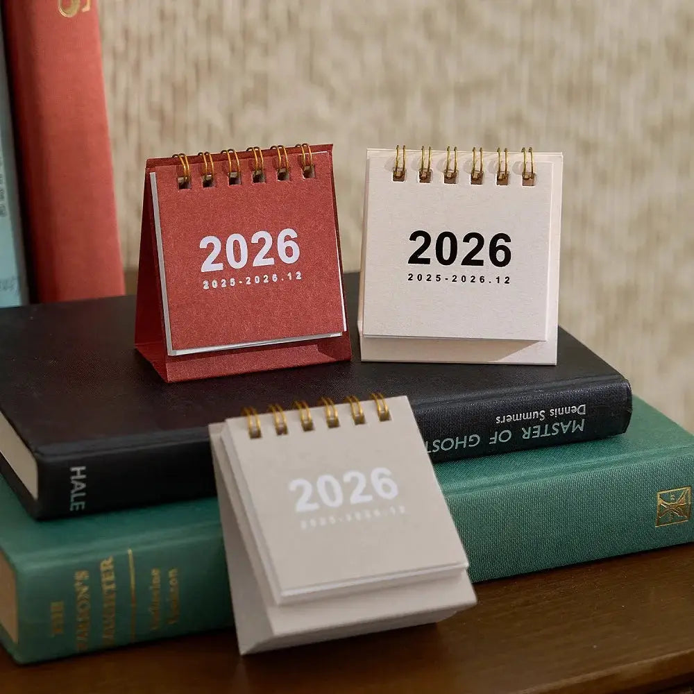 Three 2026 mini desk calendars with spiral bindings in red, beige and gray, displayed on stacked books.