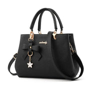 Black textured faux-leather handbag with dual top handles, crossbody strap, bow charm, and gold hardware