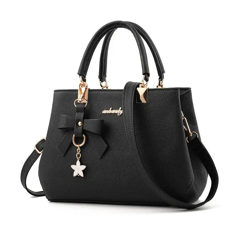 Black textured faux-leather handbag with dual top handles, crossbody strap, bow charm, and gold hardware