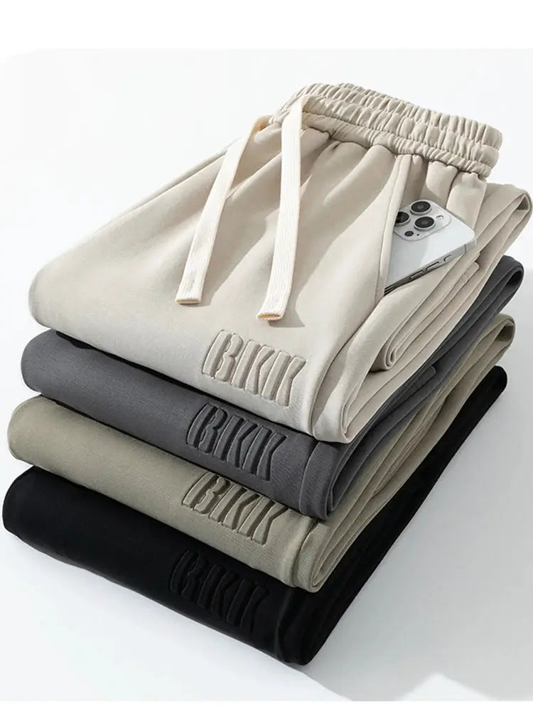 Men's wide-leg cotton track pants, 5-pair stack in beige, gray, taupe, olive, and black with drawstrings.