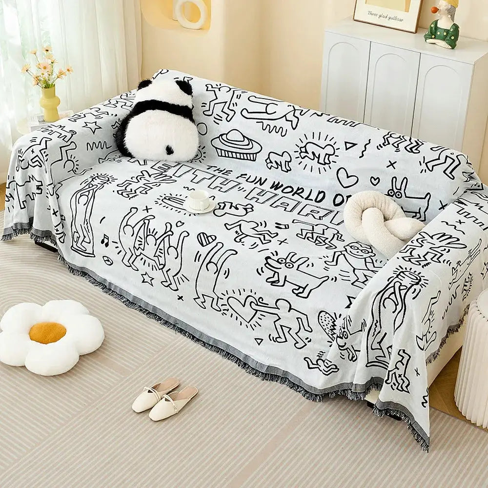Cream sofa throw blanket with black line-drawn doodle print and fringe trim draped on a couch in a cozy living room.