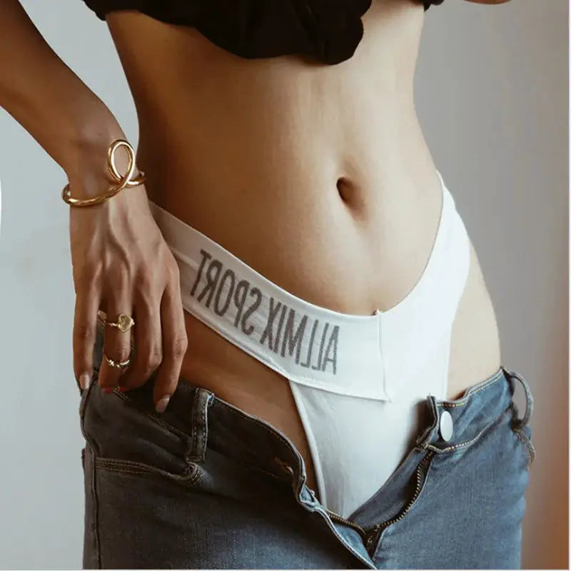 White thong underwear with elastic wide waistband and letter-print band, worn above unbuttoned denim jeans.