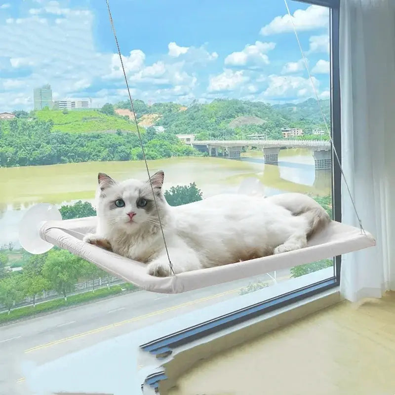 Cat hanging hammock bed, cream color, suspended by cords; cat relaxing on sunny window seat with river view.