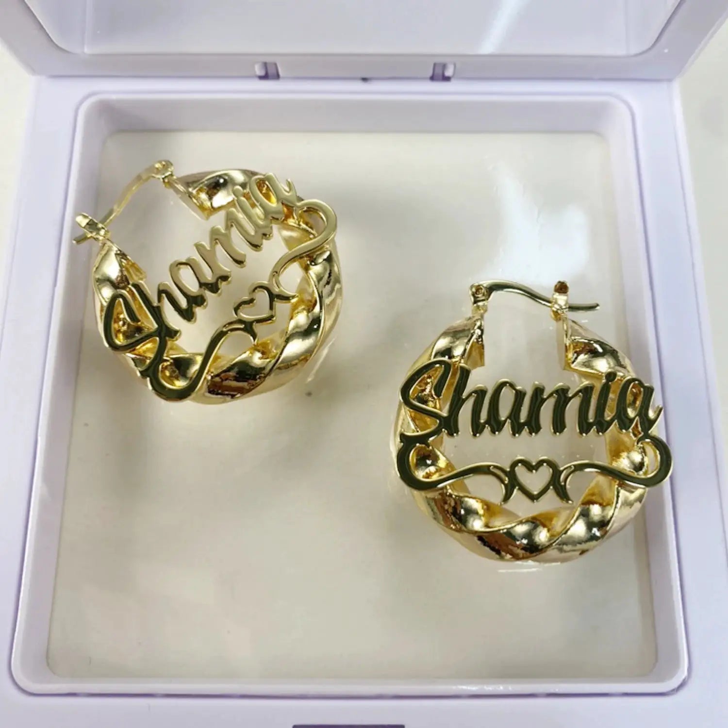 Pair of gold-tone name hoop earrings with Shania script and heart details, displayed in a jewelry box.