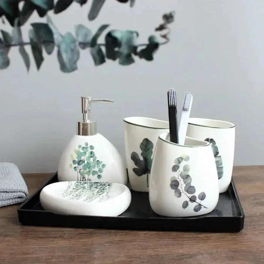 5-piece white ceramic bathroom accessory set on a black tray with soap dispenser, toothbrush holder, mouthwash cup, and matching soap dish with green botanical prints.