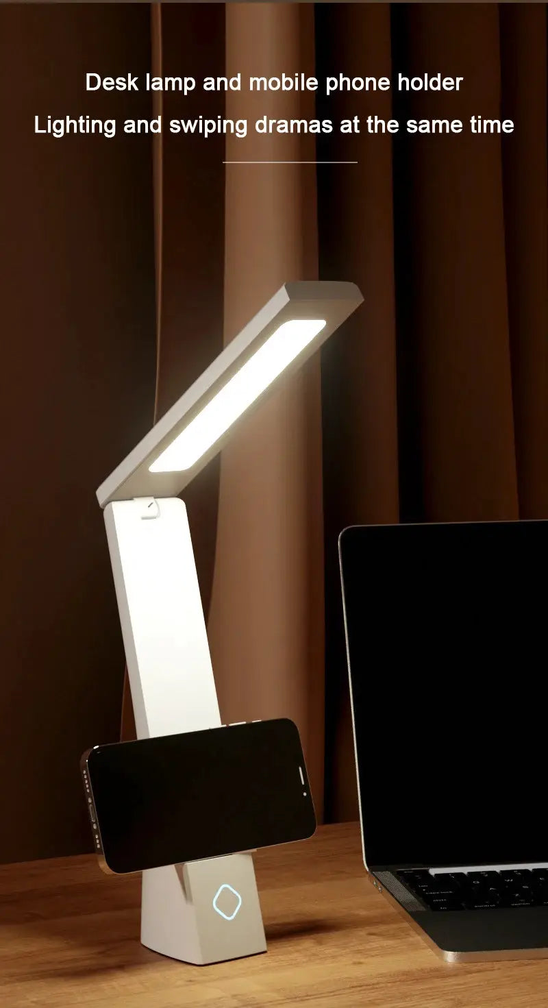 Foldable white LED desk lamp with phone stand and charging feature, resting on a wooden desk beside a laptop.