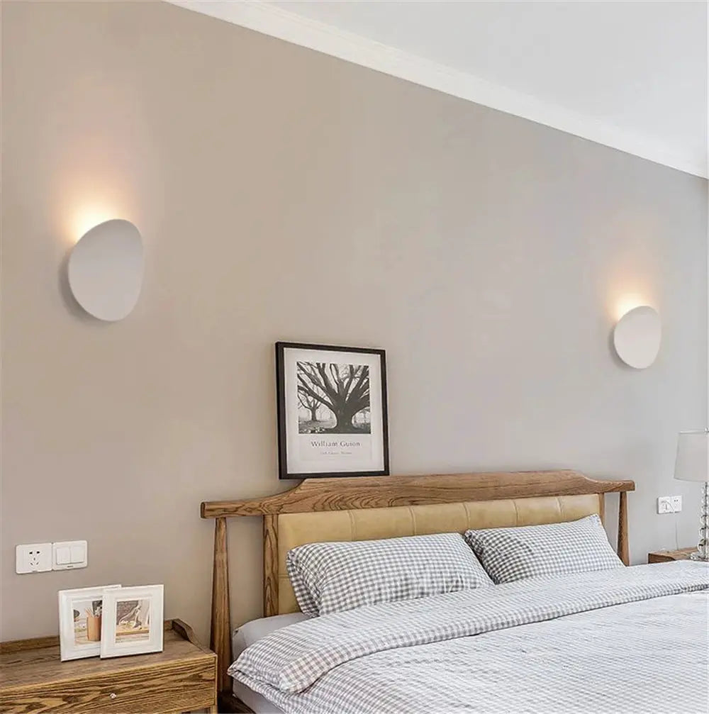 Set of two white round LED indoor wall lamps on a beige bedroom wall above a wooden bed, emitting warm light.
