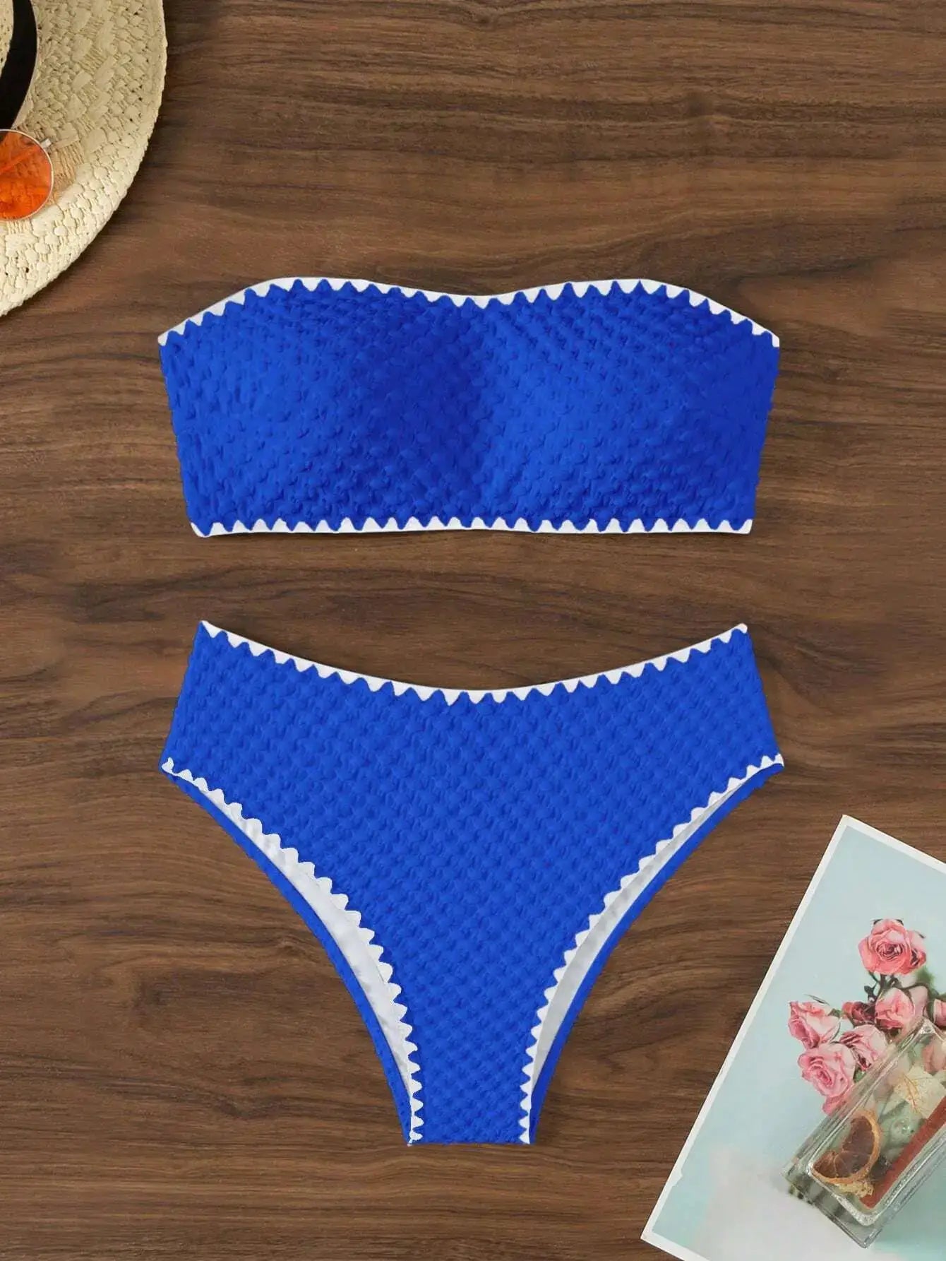 Blue strapless bandeau bikini set with white zigzag trim; top and high-cut bottoms on a wooden table.