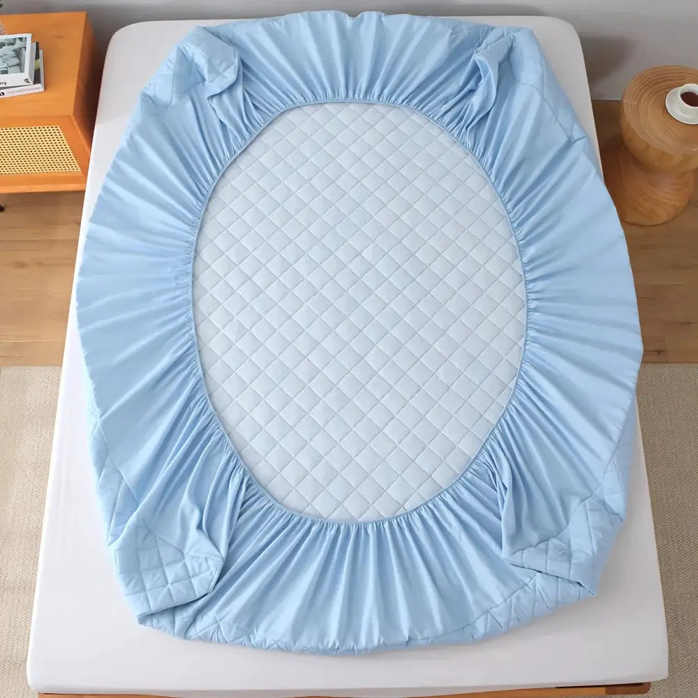Quilted waterproof fitted sheet protector in light blue with a grey diamond-quilted center on a bed