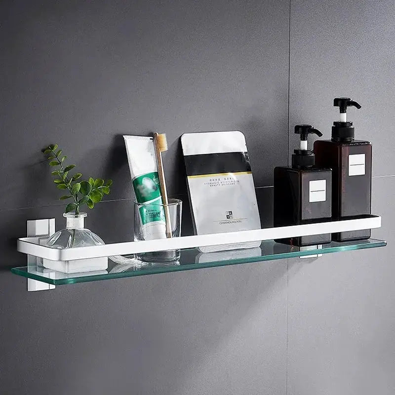 Glass bathroom shelf with white wall-mount bracket, displaying plant, toothbrush in glass, sachet, and two pump bottles
