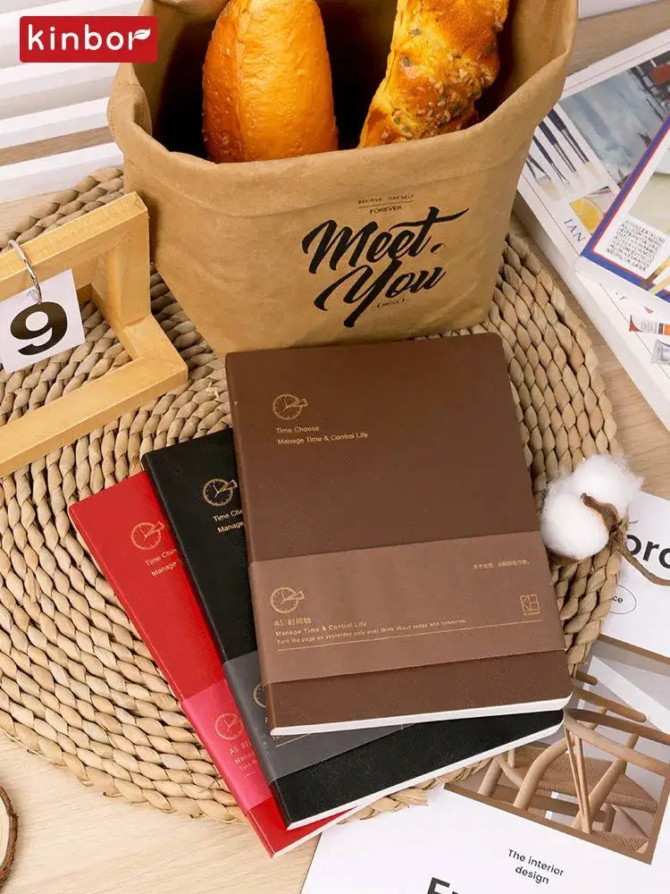 Kinbor weekly planner notebooks in brown, red, and black covers stacked on woven mat; Kinbor logo on a paper bag.