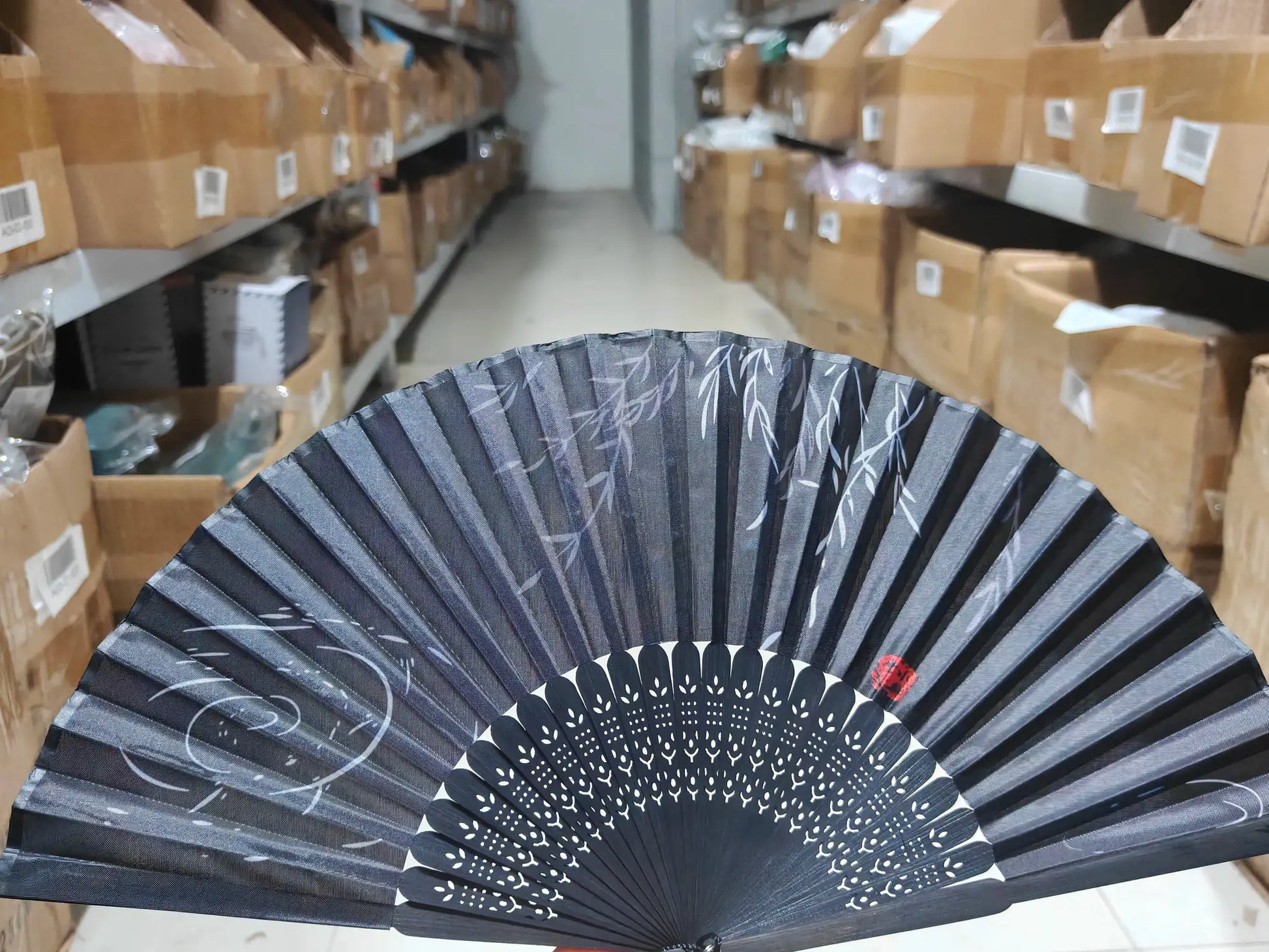 1pc black bamboo hand folding fan with white leaf pattern, shown in a warehouse aisle