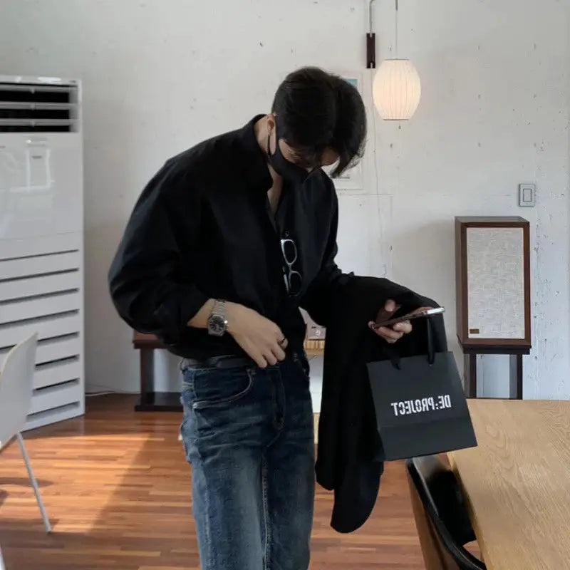 Men's black long-sleeve shirt with blue jeans, checking his phone while holding a coat and a shopping bag.