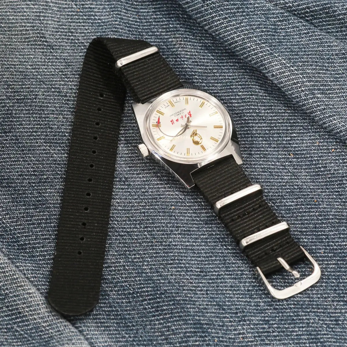 Men's 36mm hand-wind mechanical watch with silver case, white dial, gold baton markers, black nylon NATO strap.