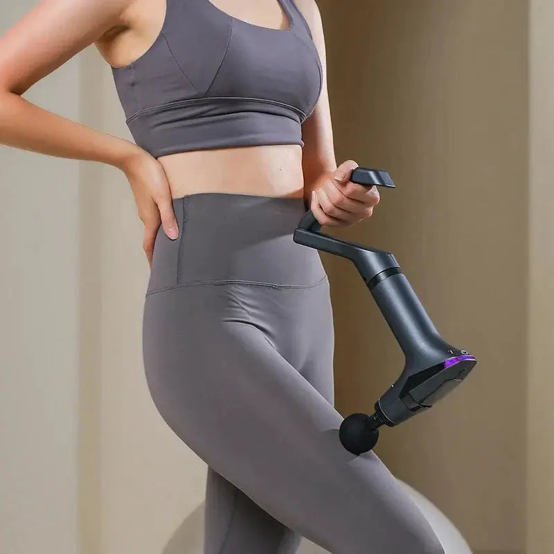 Long-handle electric massage gun, black with purple accents, held by a woman in gray workout wear