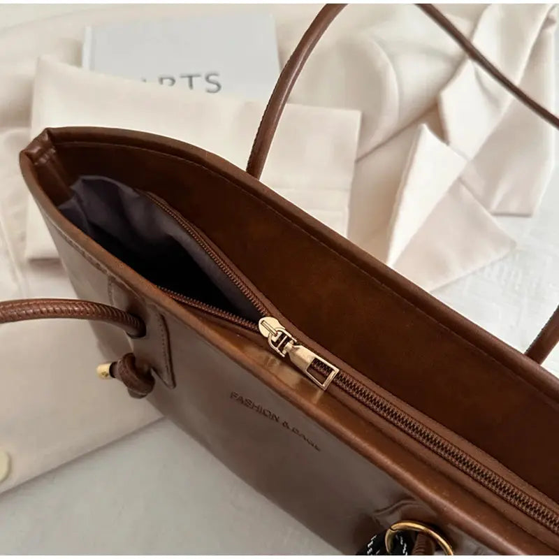 Brown faux leather tote bag with a gold top zipper, long handles, and gray interior lining