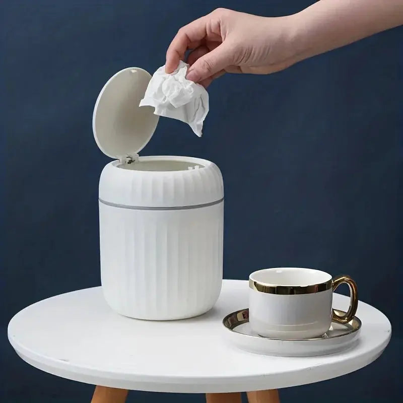 Mini white ribbed plastic desktop trash can with lid on a round table beside a teacup.
