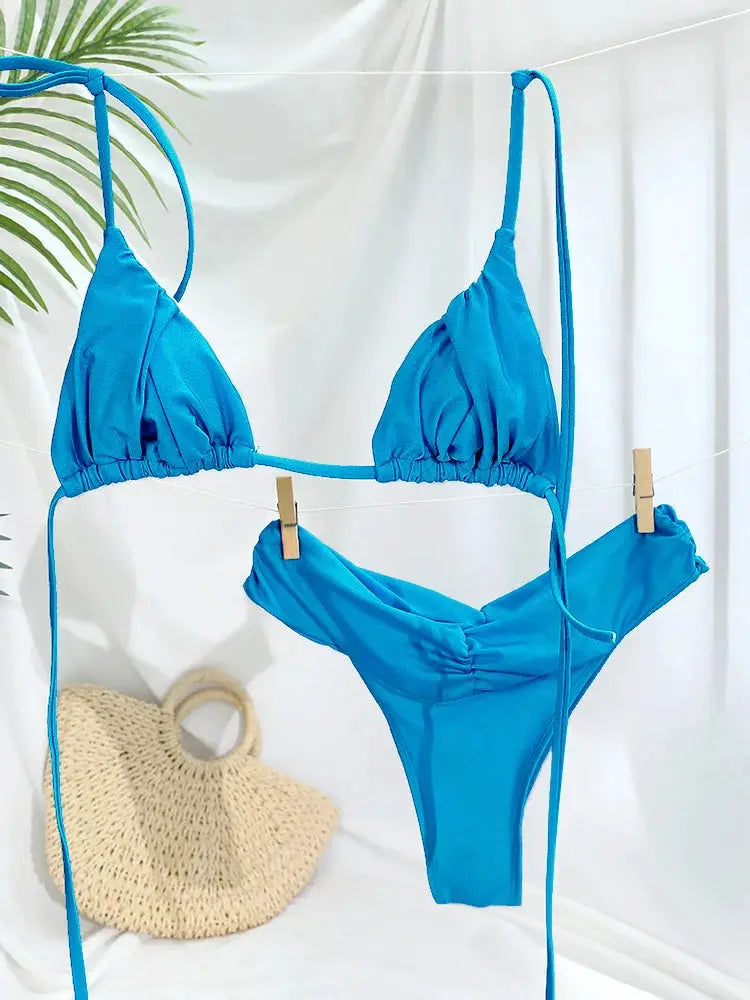Bikini set in blue with triangle cups and string ties, hanging on a clothesline with a straw bag nearby.