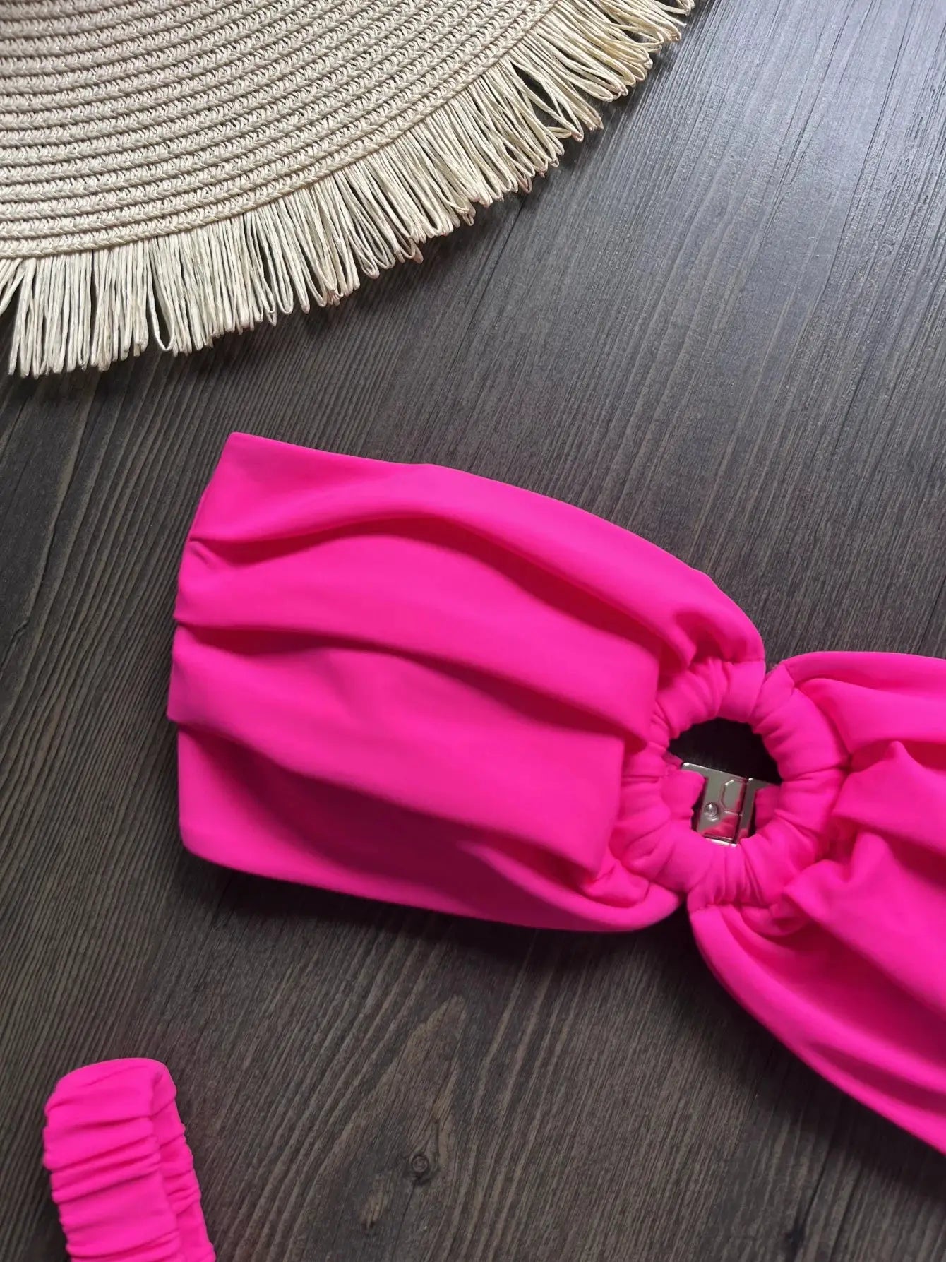 Pink micro bikini top with ruched bandeau and metal clasp, on dark wood with a straw-fringed hat nearby.