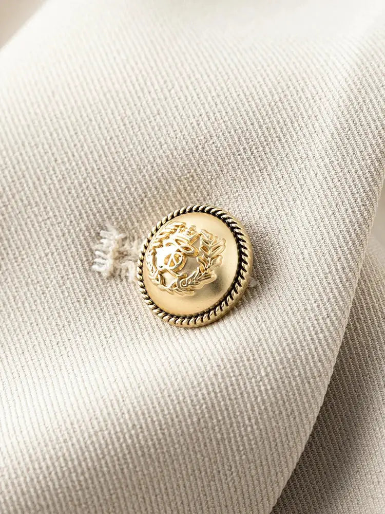 Beige blazer fabric close-up with gold-tone ornate button and twisted edge.