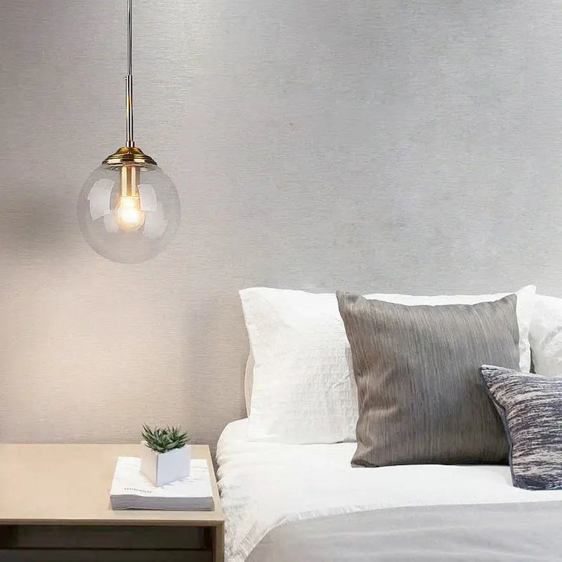 Modern luxury metal pendant lamp with glass globe and brass top, single hanging fixture over a bed.