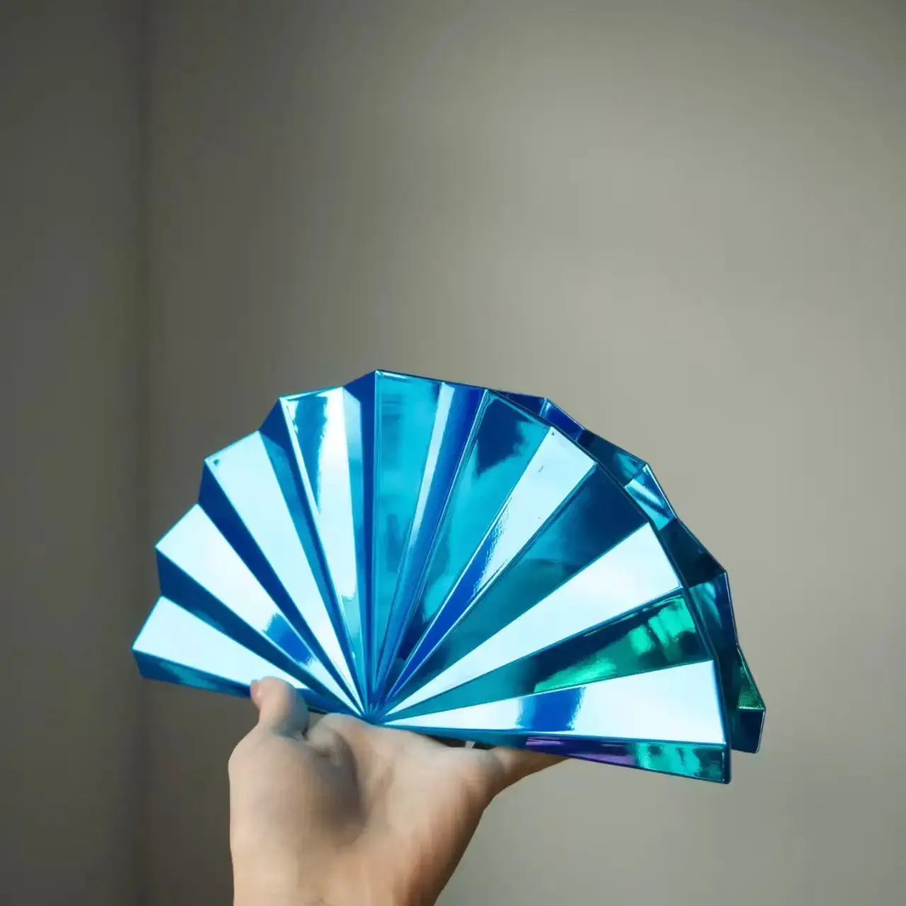 Blue metallic folding hand fan held by a hand against a neutral background.